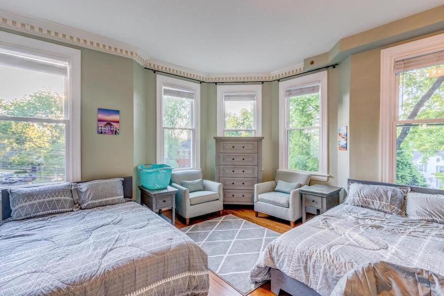 Bedroom with two beds, gray bedding, armchairs, chest of drawers, large windows, and a green wall with artwork.