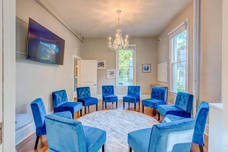 Room with blue velvet chairs arranged in a circle, chandelier, and large windows with a wall-mounted TV.