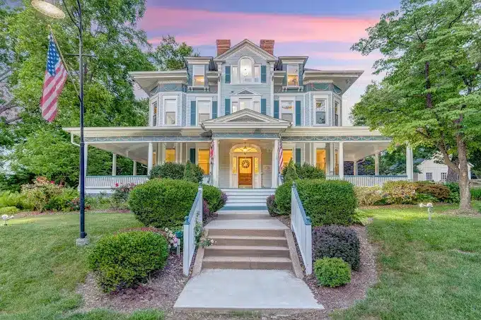 Victorian-style house with wraparound porch, manicured garden, and evening sky background.