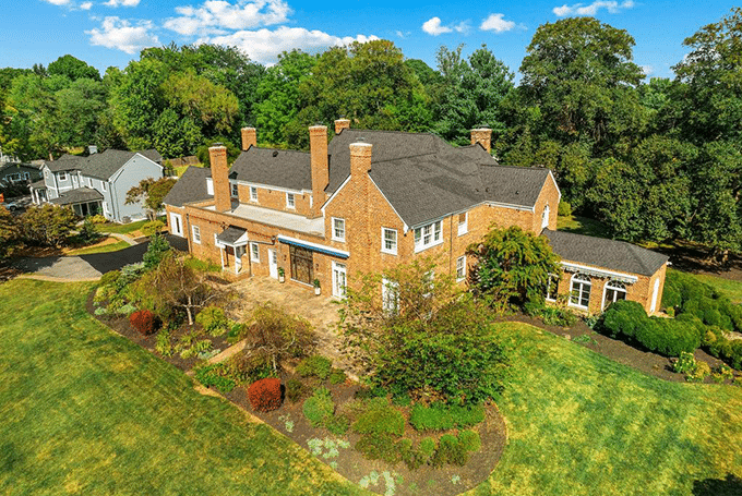 Aerial view of a large brick house with lush gardens and green lawns under a blue sky.