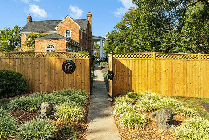 Charming brick house with a wooden fence, open gate, and garden path flanked by lion statues under a blue sky.