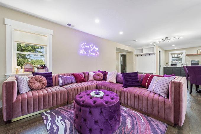 Stylish living room with pink velvet sectional sofa, purple accents, zebra rug, and neon sign reading One Day at a Time.