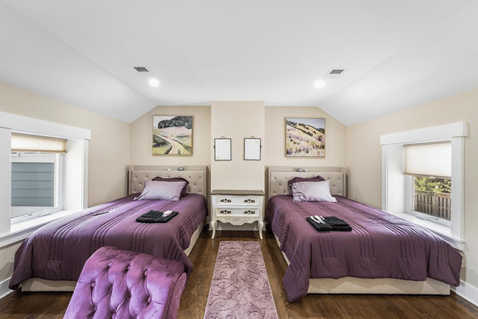 Cozy bedroom with two purple-accented twin beds, white nightstand, and artwork on beige walls.