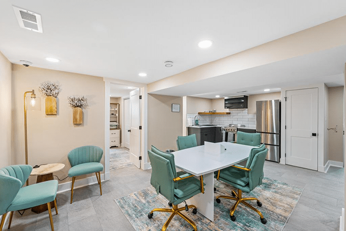 Modern home office with teal chairs and white table, kitchen in background, bright lighting, and neutral walls.