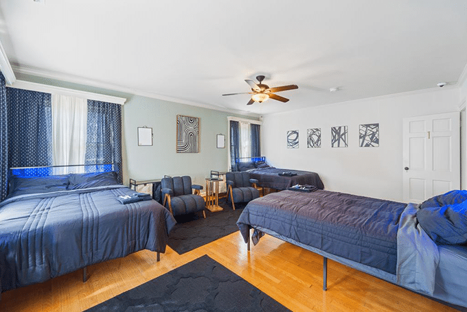 Spacious bedroom with three beds, modern decor, ceiling fan, and natural light from large windows.